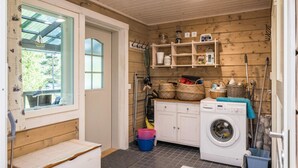 Een wasserette met een wasmachine, planken met manden, een bankje en een raam met uitzicht op groen.