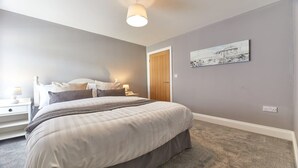 A bedroom with a bed, nightstands, a lamp, a painting, and a door. - Host & Stay | Sea Breeze (Amble)