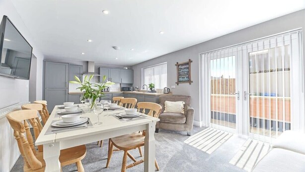 A modern kitchen with grey cabinets, a dining area with a white table set for four, and a sliding glass door leading to an outdoor patio. - Host & Stay | Sea Breeze (Amble)