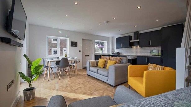 Uma sala de estar moderna com sofá, área de refeições com cadeiras, cozinha com armários e uma televisão na parede.