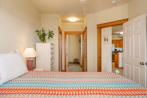 A bedroom with a bed, a nightstand, a lamp, a dresser, and a potted plant. There is a hallway leading to a kitchen area.