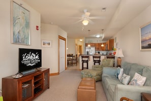A living room with a television, a ceiling fan, a couch, and a dining area with chairs and a table. - La Dolce Vita II: Soundfront condo w/sunset views, community pool, 2 en-suites! (Nags Head)