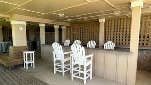 A covered outdoor bar area with a wooden deck, white Adirondack chairs, and a clock on a pillar. - La Dolce Vita II: Soundfront condo w/sunset views, community pool, 2 en-suites! (Nags Head)