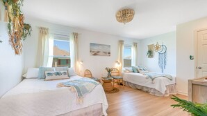 Una habitación con dos camas, una mesita pequeña y una silla. Se ve una ventana con cortinas, una obra de arte en la pared y una lámpara en el techo.