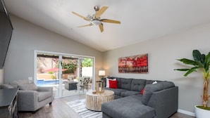 A modern living room with a grey sectional sofa, a round wooden coffee table, a ceiling fan, and a large potted plant. - Casa De Mojave 3 Bedroom Home by RedAwning (Scottsdale)