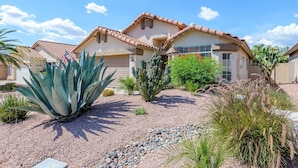 A two-story house with a tiled roof, a garage, and a well-maintained garden with various plants and a large agave plant. - Casa De Mojave 3 Bedroom Home by RedAwning (Scottsdale)