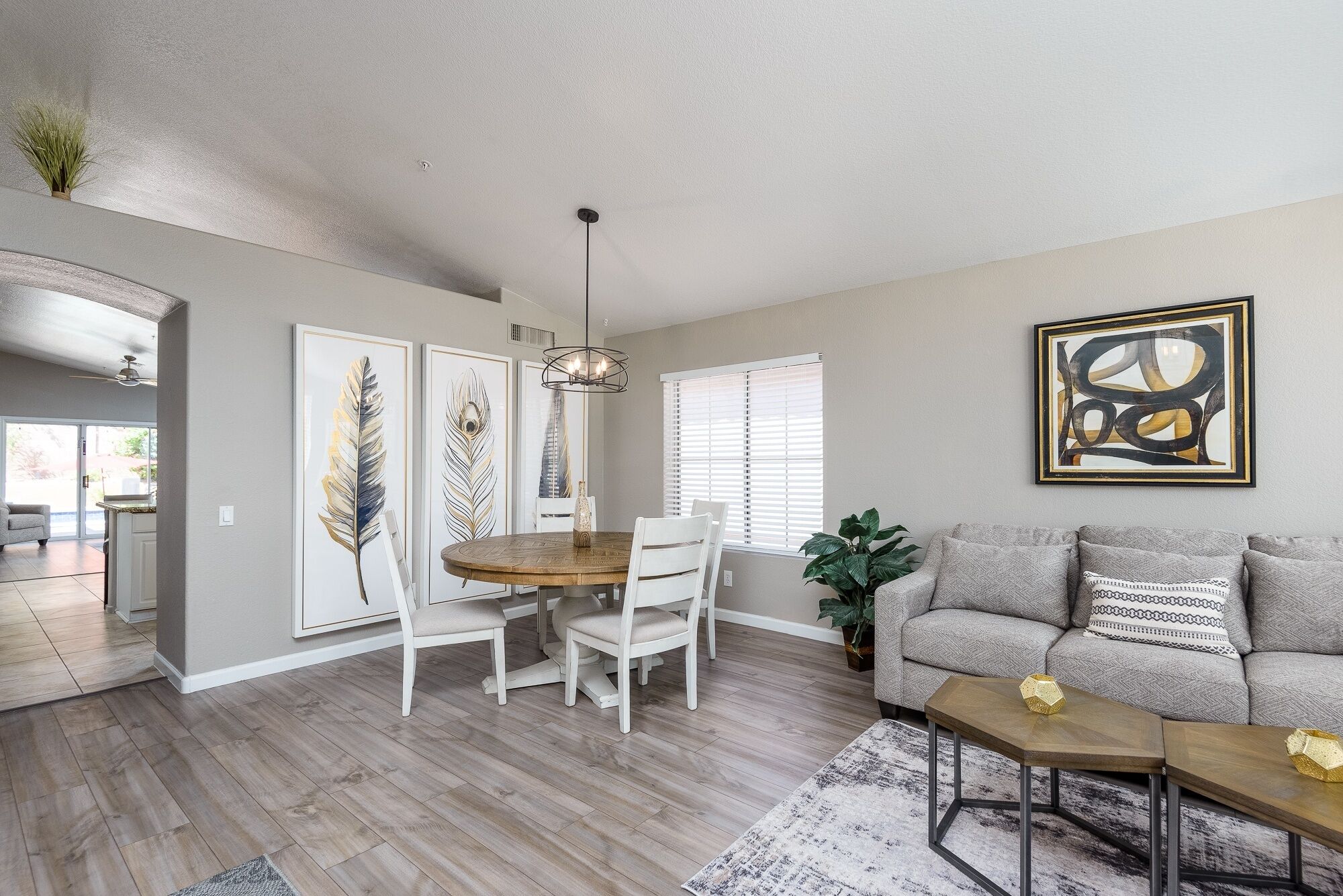 A modern living room with a dining area, featuring a round wooden table, white chairs, a grey sofa, and a large abstract painting.