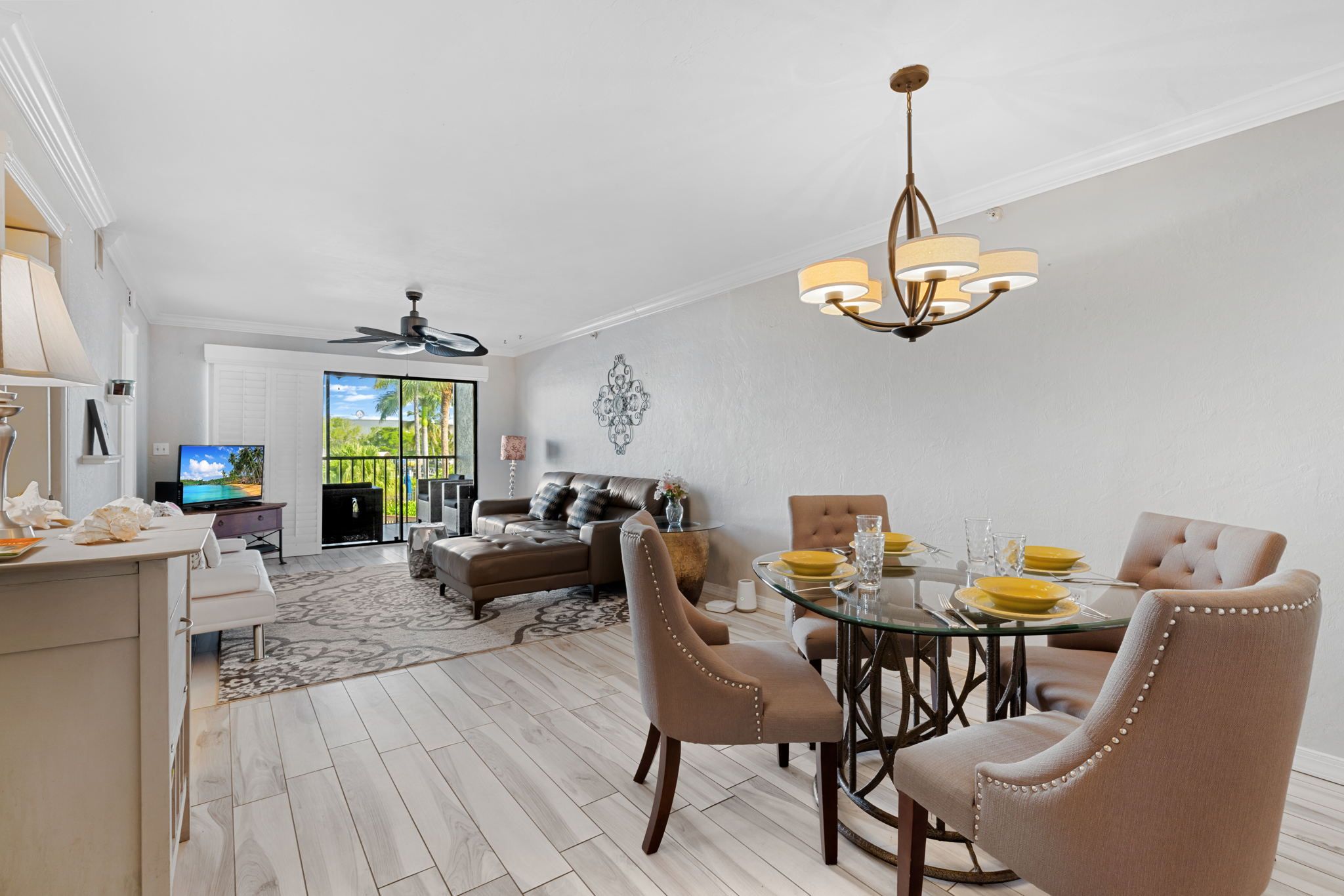 A modern living room with a dining area, featuring a glass table, beige upholstered chairs, a ceiling fan, and a television on a white cabinet.