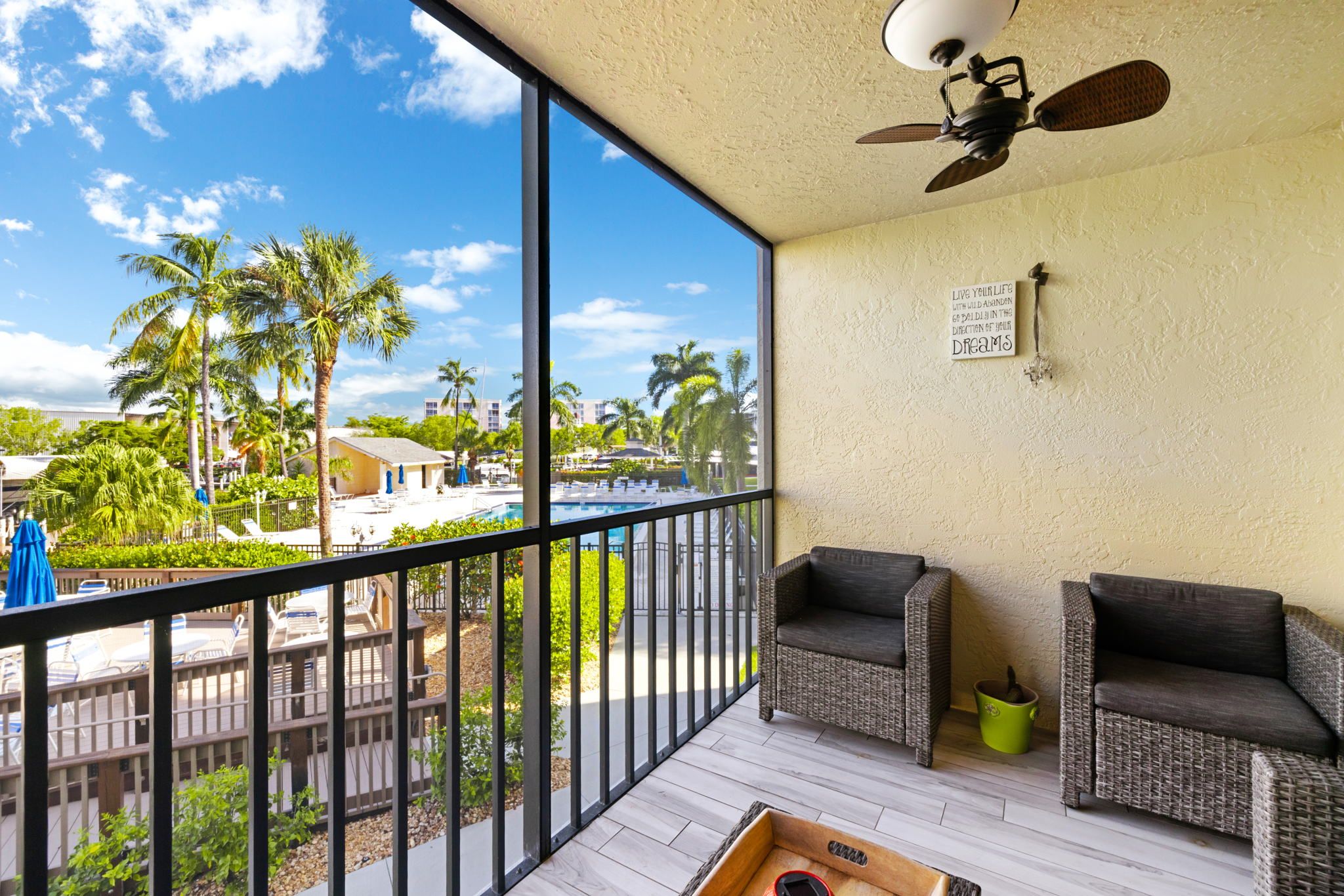 A balcony with wicker furniture, a ceiling fan, and a view of a pool and palm trees.