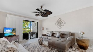 A modern living room with a sofa, ottoman, and coffee table, a ceiling fan, and a television on a stand. - Santa Maria II 110 by Distinctive Beach Rentals (Fort Myers Beach)