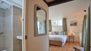 Une chambre avec un plancher en bois, un lit recouvert d’une couette blanche, une table de chevet avec une lampe, un miroir et une fenêtre avec des rideaux.
