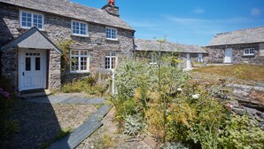 Une petite maison en pierre avec une porte et des fenêtres blanches, entourée d’un jardin où poussent différentes plantes et bordée d’un chemin en pierre.