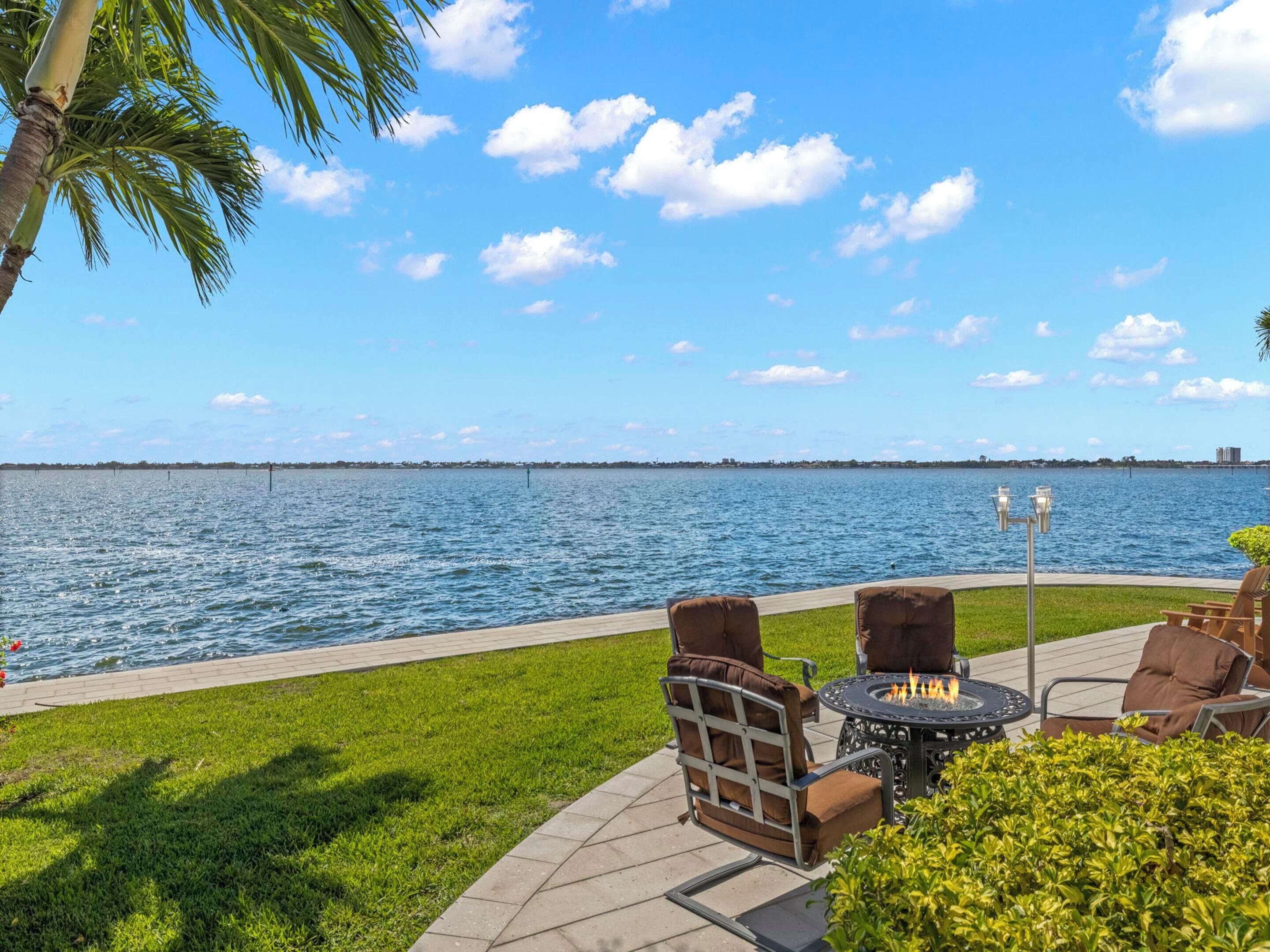 An outdoor patio with a fire pit table and chairs, overlooking a body of water with a distant shoreline.