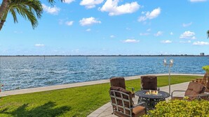 An outdoor patio with a fire pit table and chairs, overlooking a body of water with a distant shoreline. - Dolce Vita Villa by Distinctive Beach Rentals (Cape Coral)