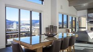 A modern dining area with a glass table and chairs, large windows with a view of mountains and a city, and a cozy seating area with a sofa and TV. - Rendezvous Game Trail | Luxe Mountain Home | Brand New Build | Amazing Views (Fraser)
