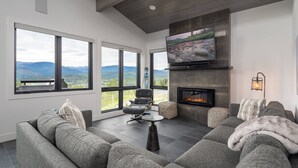 A modern living room with a large gray sectional sofa, a small round coffee table, and a fireplace. There are two large windows offering a view of mountains. - Rendezvous Game Trail | Luxe Mountain Home | Brand New Build | Amazing Views (Fraser)