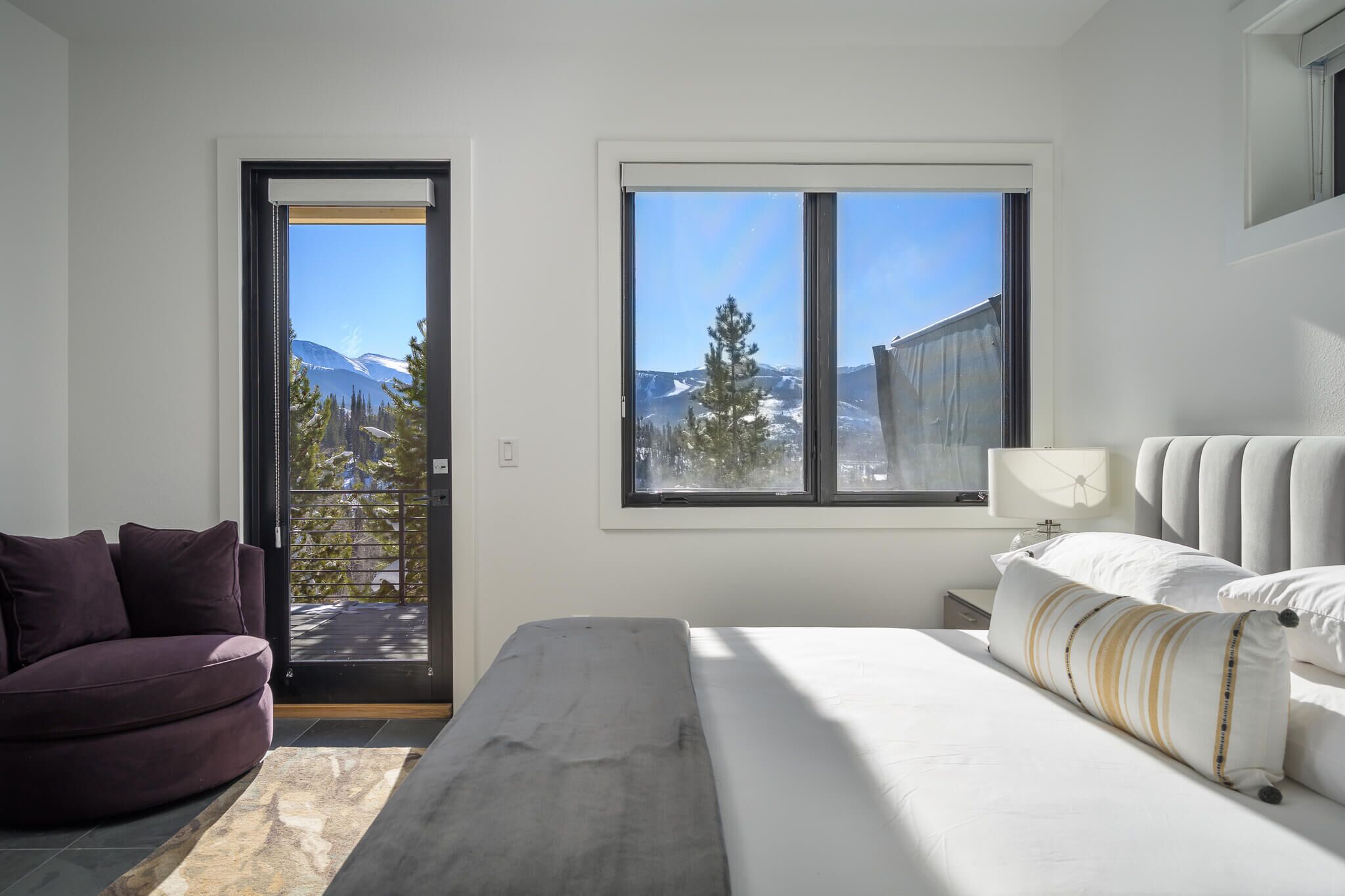A bedroom with a bed, a sofa, a window with a view of mountains, and a door leading outside.