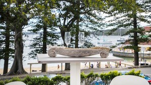 Une table ornée d’un objet décoratif et de deux verres, donnant sur un front de mer où l’on aperçoit des personnes et des bateaux.