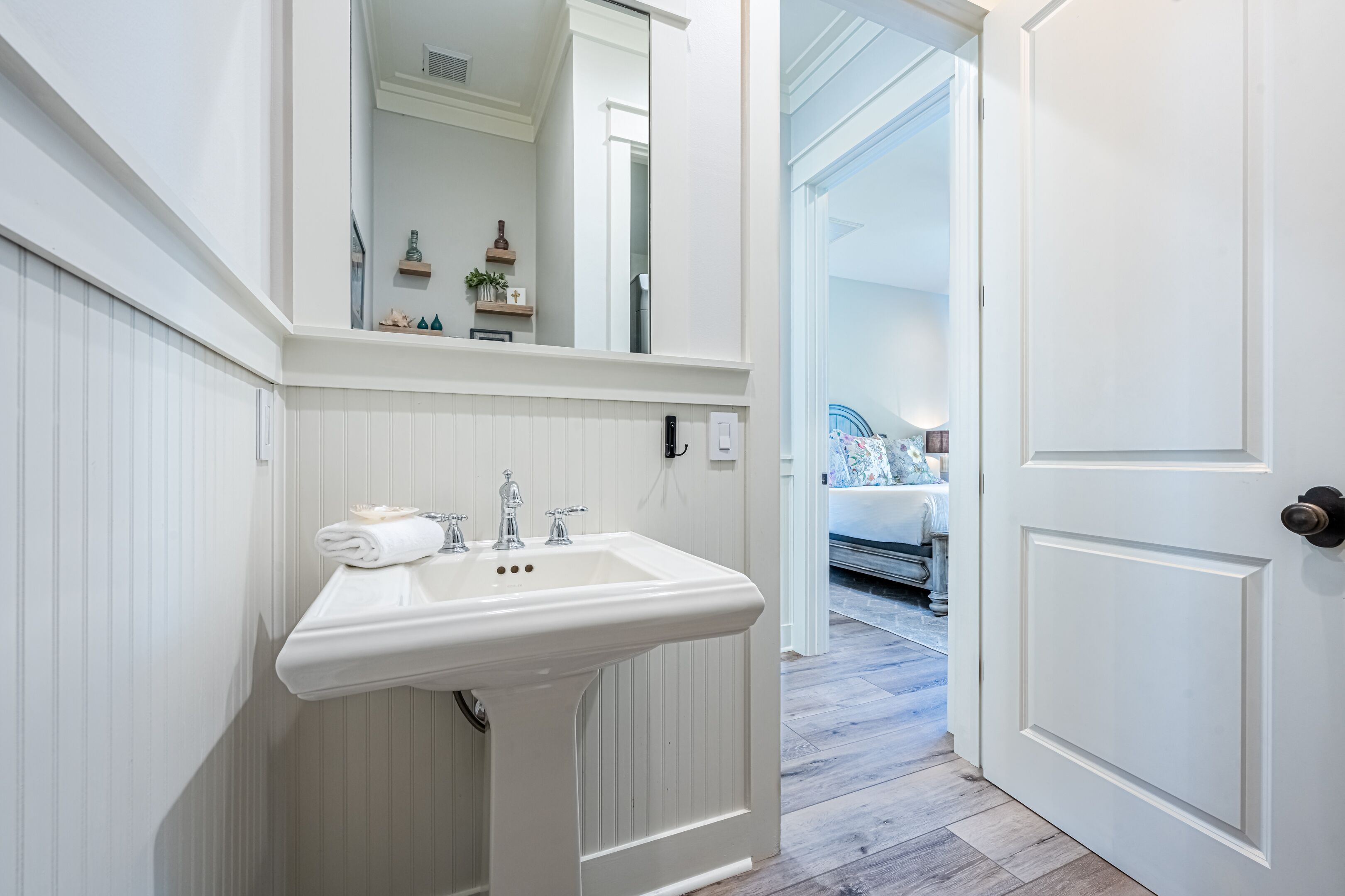 Un baño con lavamanos blanco, espejo y puerta que da a un dormitorio con cama y mesita de noche.