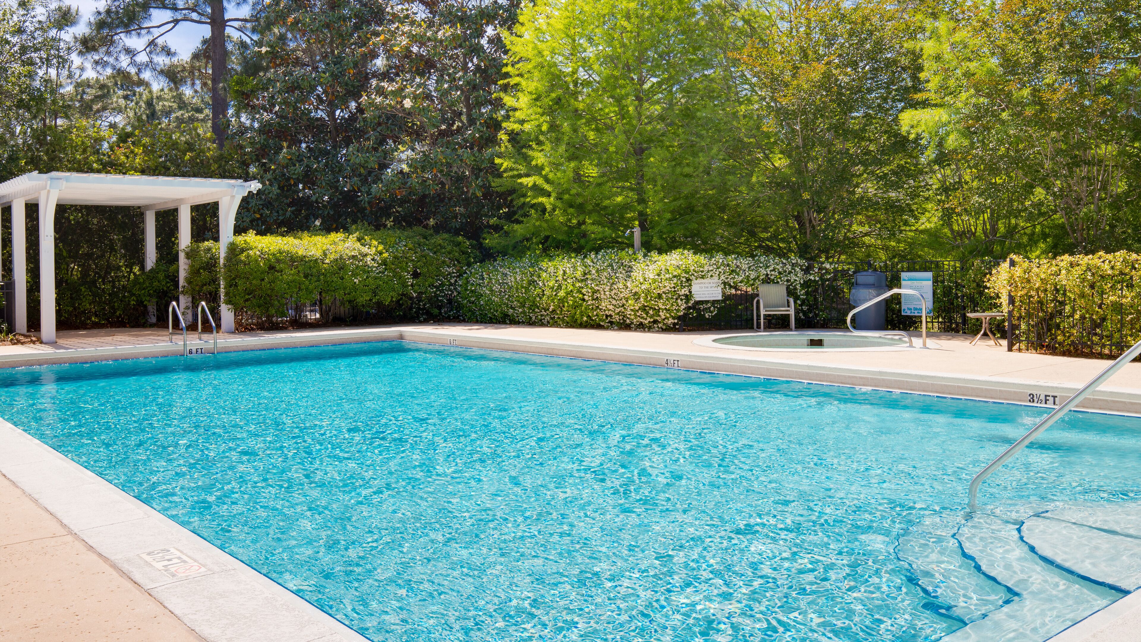 Een rechthoekig zwembad met helderblauw water, omgeven door een wit terras en een overdekt zitgedeelte.