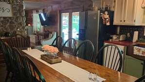 Une table à manger en bois, avec des chaises, un lustre et un coin cuisine équipé de placards et d’un réfrigérateur.