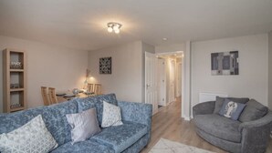 A living room with a blue sofa, a grey armchair, a dining table with chairs, and a hallway leading to another room.
