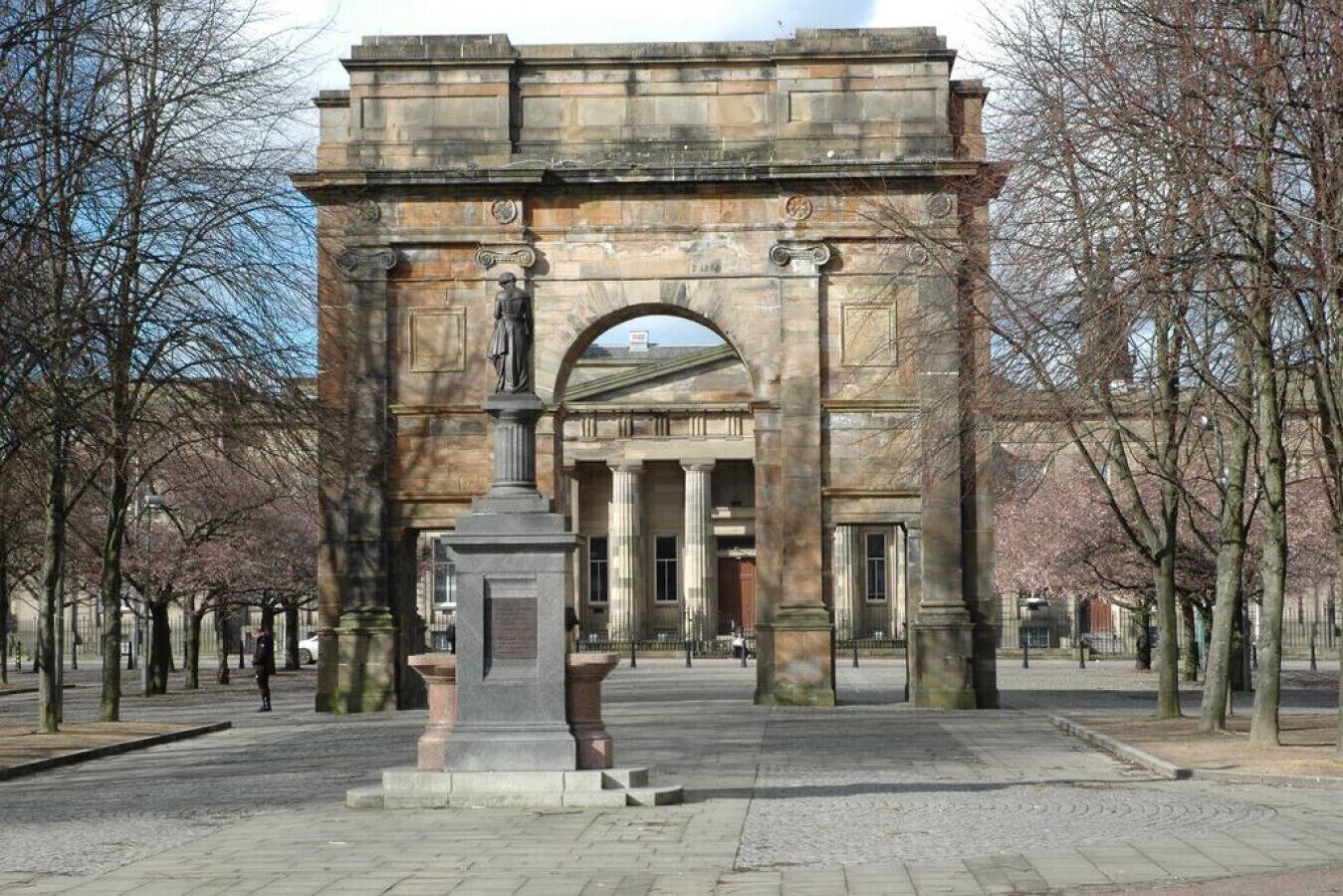 A stone archway with a statue in the center, surrounded by trees and a building in the background.