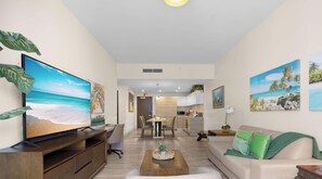 A modern living room with a large flat-screen TV, a wooden coffee table, a beige sofa with green cushions, and a dining area with a kitchen in the background.