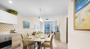 A modern kitchen with white cabinets, a dining area with a long table, and a beach-themed painting on the wall.