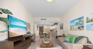 A modern living room with a large flat-screen TV, a wooden coffee table, a beige sofa with green cushions, and a dining area with a kitchen in the background.
