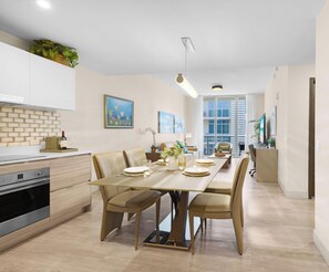 A modern kitchen with white cabinets, a dining area with a long table, and a beach-themed painting on the wall.