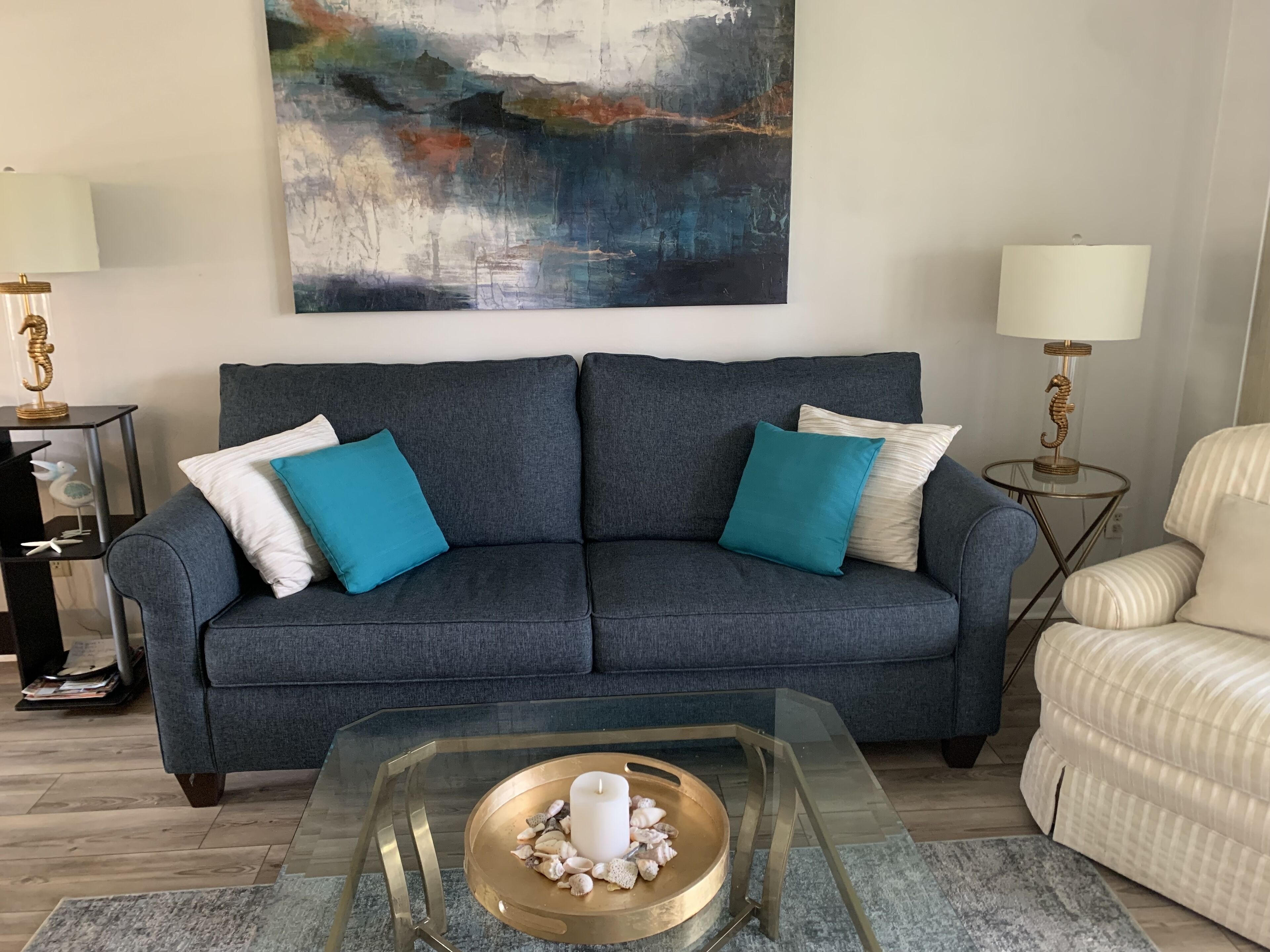 A modern living room with a grey sofa, teal cushions, a glass coffee table with a decorative centerpiece, and a large abstract painting on the wall.