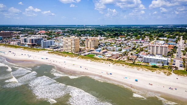 Une vue aérienne d’une ville côtière, avec une vaste plage, plusieurs immeubles de grande hauteur et un ciel bleu dégagé.