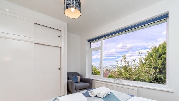 Ett sovrum med en säng, ett fönster med utsikt över grönområden, en stol och en hängande lampa.