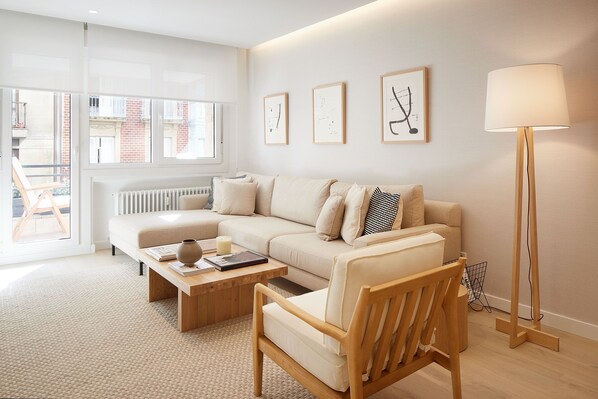 Una sala moderna con un sofá, sillones, una mesita de centro y una lámpara de pie. Se observan tres obras de arte enmarcadas en la pared y una ventana que proporciona luz natural.