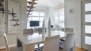 Et moderne spiseområde med et glasbord, grå polstrede stole og en trappe, der fører til en etage over.