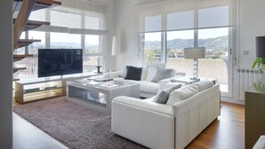 En moderne stue med en hvid sofa, et sofabord og et fjernsyn. Store vinduer med rullegardiner, en trappe og udsigt over byen.