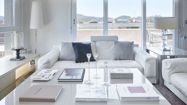 En moderne, lys stue med en hvid sofa, et sofabord med bøger og glas, samt store vinduer der giver udsigt over bybilledet.