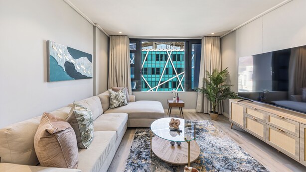 A modern living room with a sofa, a glass coffee table, a TV, and a view of a building through large windows.