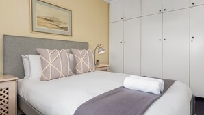 En pænt redt seng med gråt sengegavl, et natbord med en lampe, og en stor garderobe med flere døre.