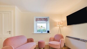 Una habitación con una puerta blanca, una ventana con vistas, un sofá rosa, un sillón rosa, un pequeño reposapiés, una lámpara de pie y un televisor montado en la pared.