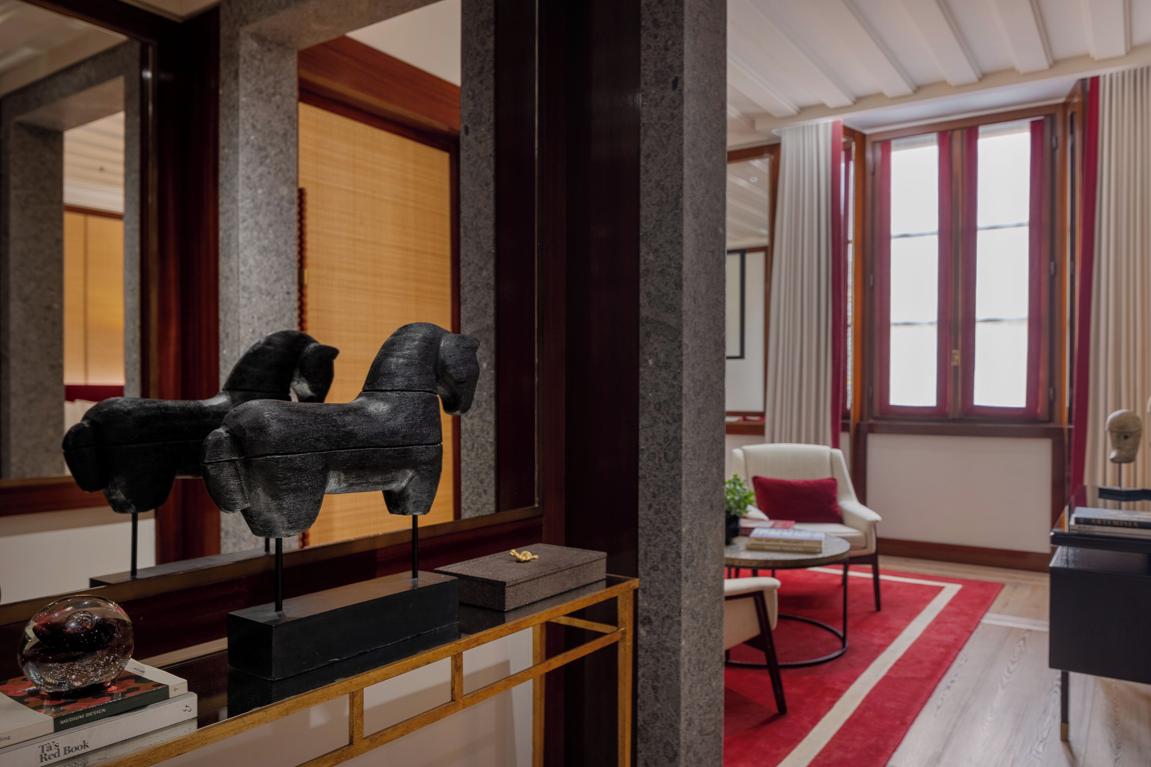 Un interior moderno con una escultura sobre una consola, una alfombra roja y vistas de una habitación tradicional a través de una ventana.