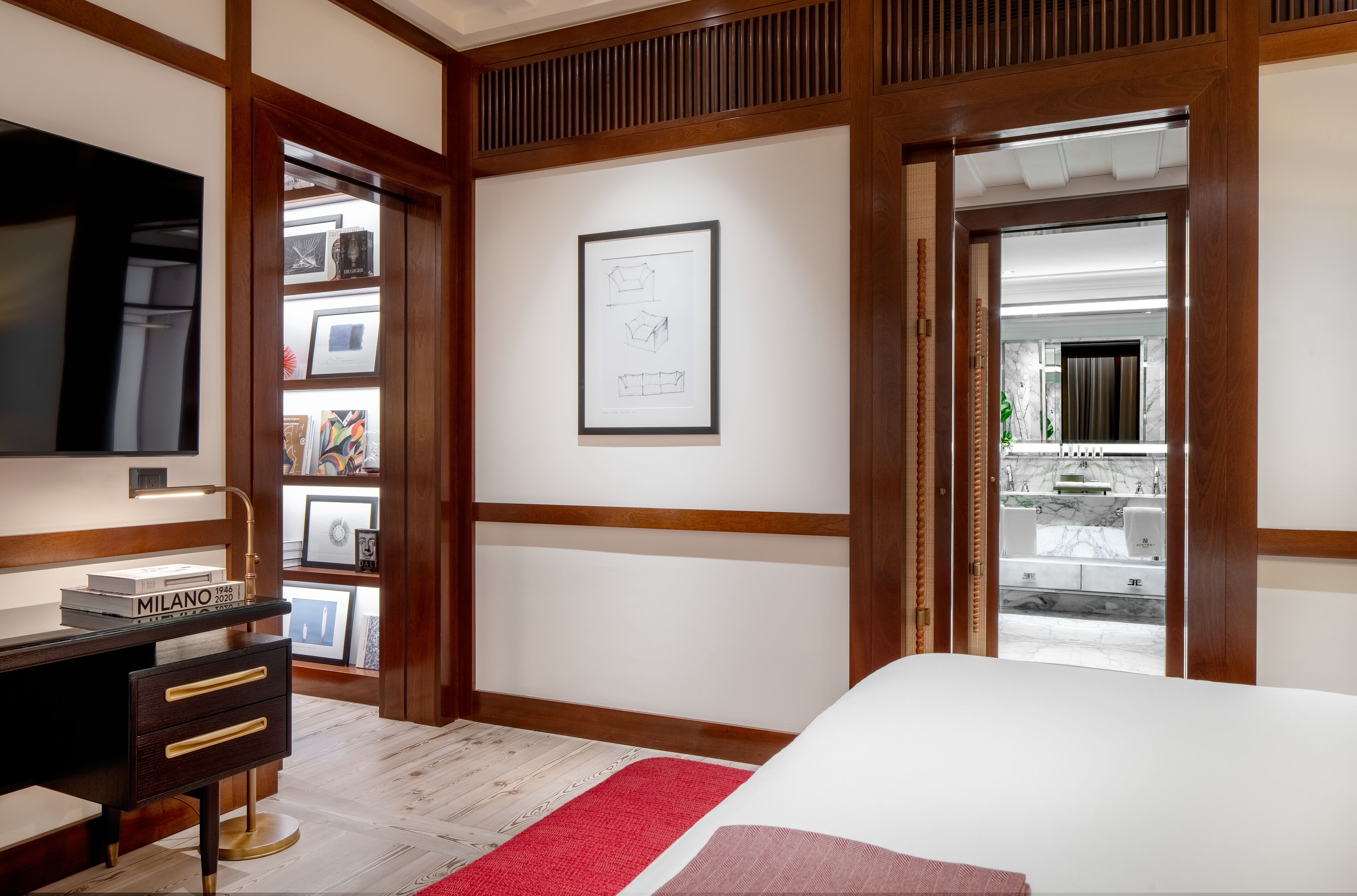 A bedroom with a bed, a desk with drawers, a framed picture on the wall, and a view of a bathroom through an open door.