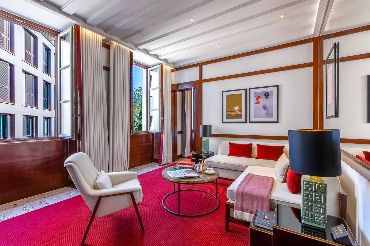 A hotel room with a red carpet, a white sofa with red cushions, a white armchair, a round coffee table, and a lamp.