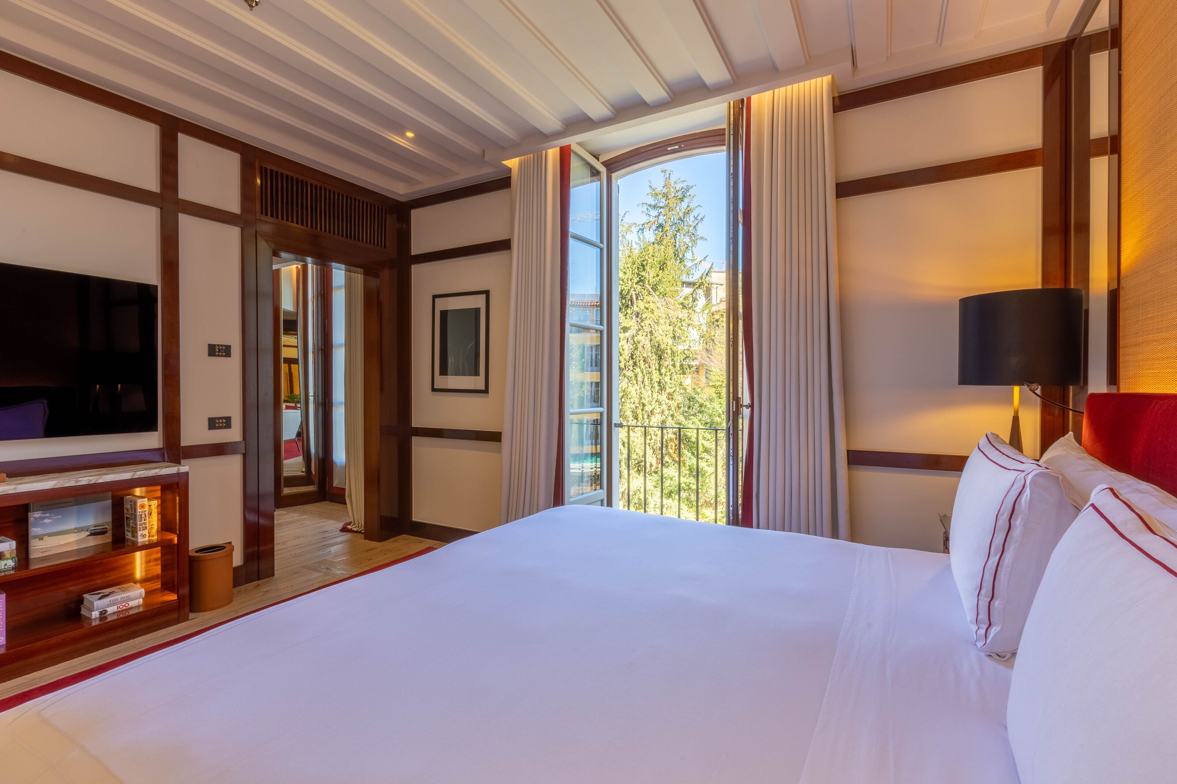 Ein Hotelzimmer mit einem großen Bett, einem Fernseher, einem Bücherregal und einem Balkon mit Blick.
