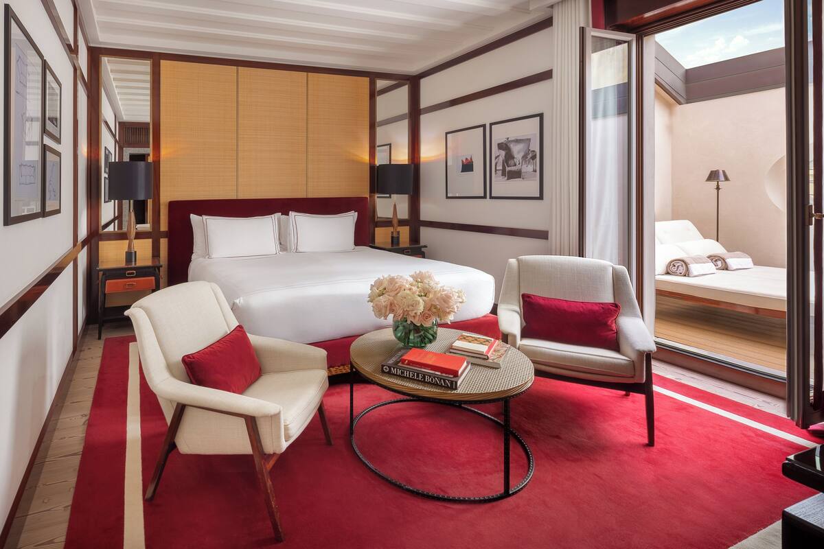 A hotel room with a large bed, two armchairs, a small round table with a book and flowers, and a view through a glass door.