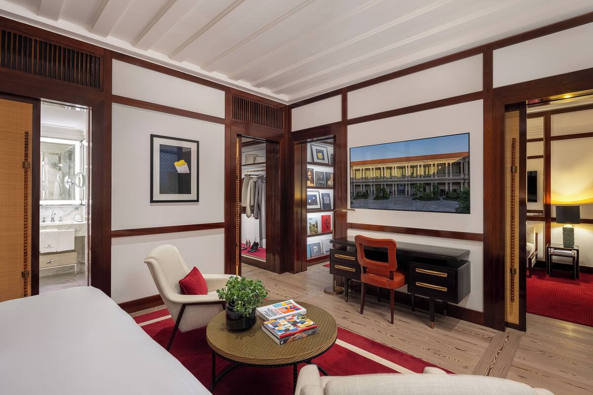 A hotel room with a large window, a fireplace, a seating area with a red rug, and a bookshelf with framed pictures.