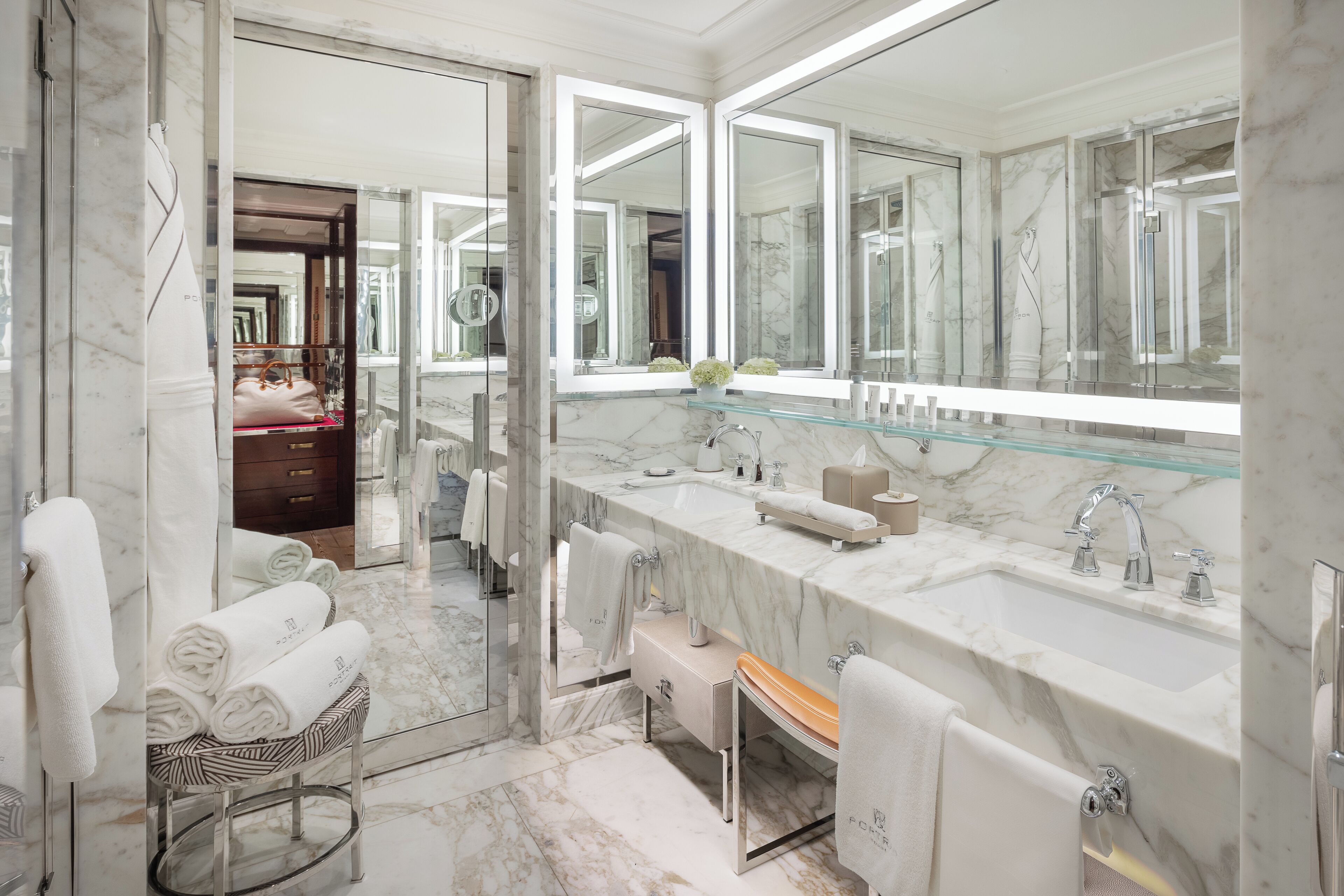 A modern bathroom with a large mirror, marble countertops, a glass shower enclosure, and a vanity with a bench.