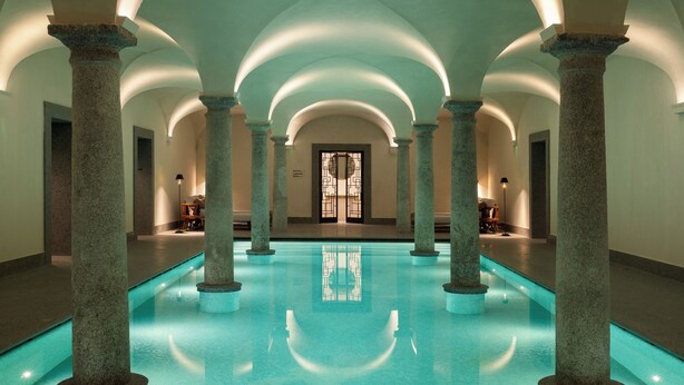 Una piscina interior amplia con columnas y arcos de piedra, una puerta con un vitral, y un ambiente tranquilo.