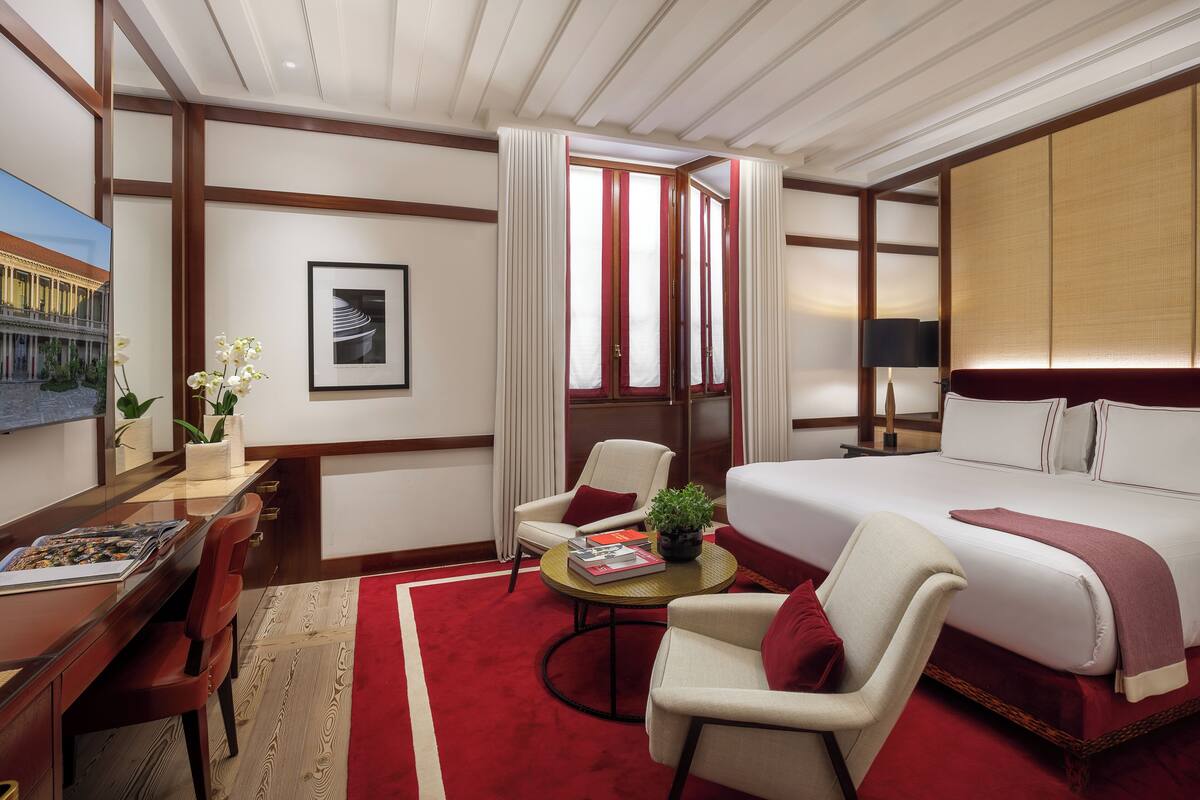 A hotel room with a large bed, a desk with a chair, a small table with a plant, and a painting on the wall.